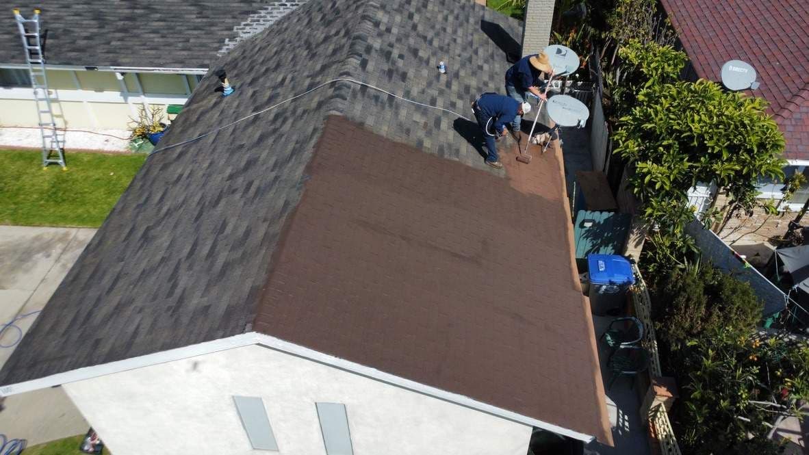 Crew rolling brown acrylic coating over gray composition shingle in Oxnard.