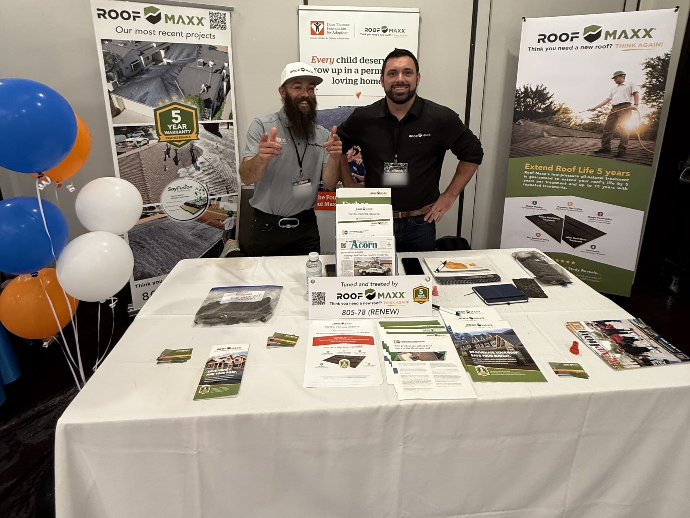 Two of our team at an expo booth beside a table of shingle samples, handouts, and a copy of the Acorn
