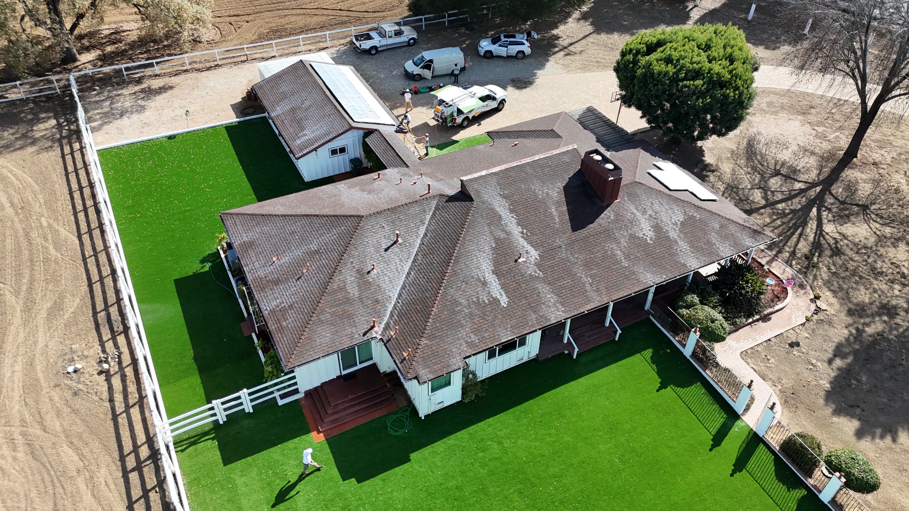 Aerial view of a long single-story estate roof mid-treatment, pale milky patches of freshly sprayed Roof Maxx across the brown composition slopes, crew working and hoses run from the truck in the drive.