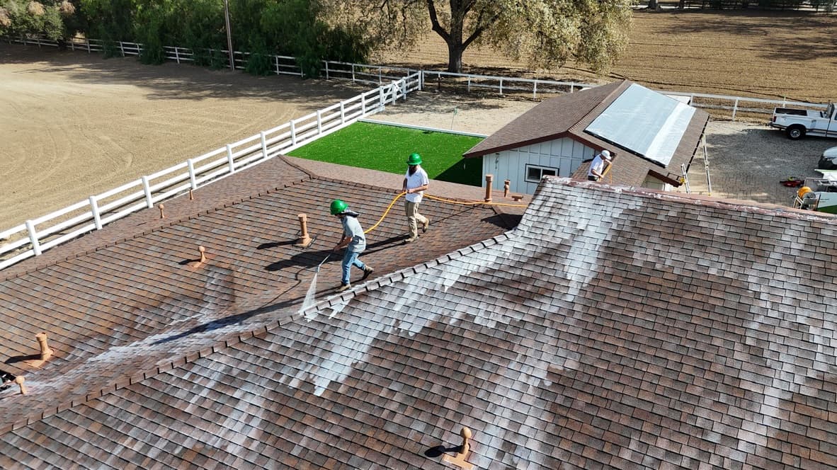 Aerial view of a large estate roof mid-treatment with three crew members working, milky white Roof Maxx spreading down the near slopes across the brown shingle, a riding arena beyond