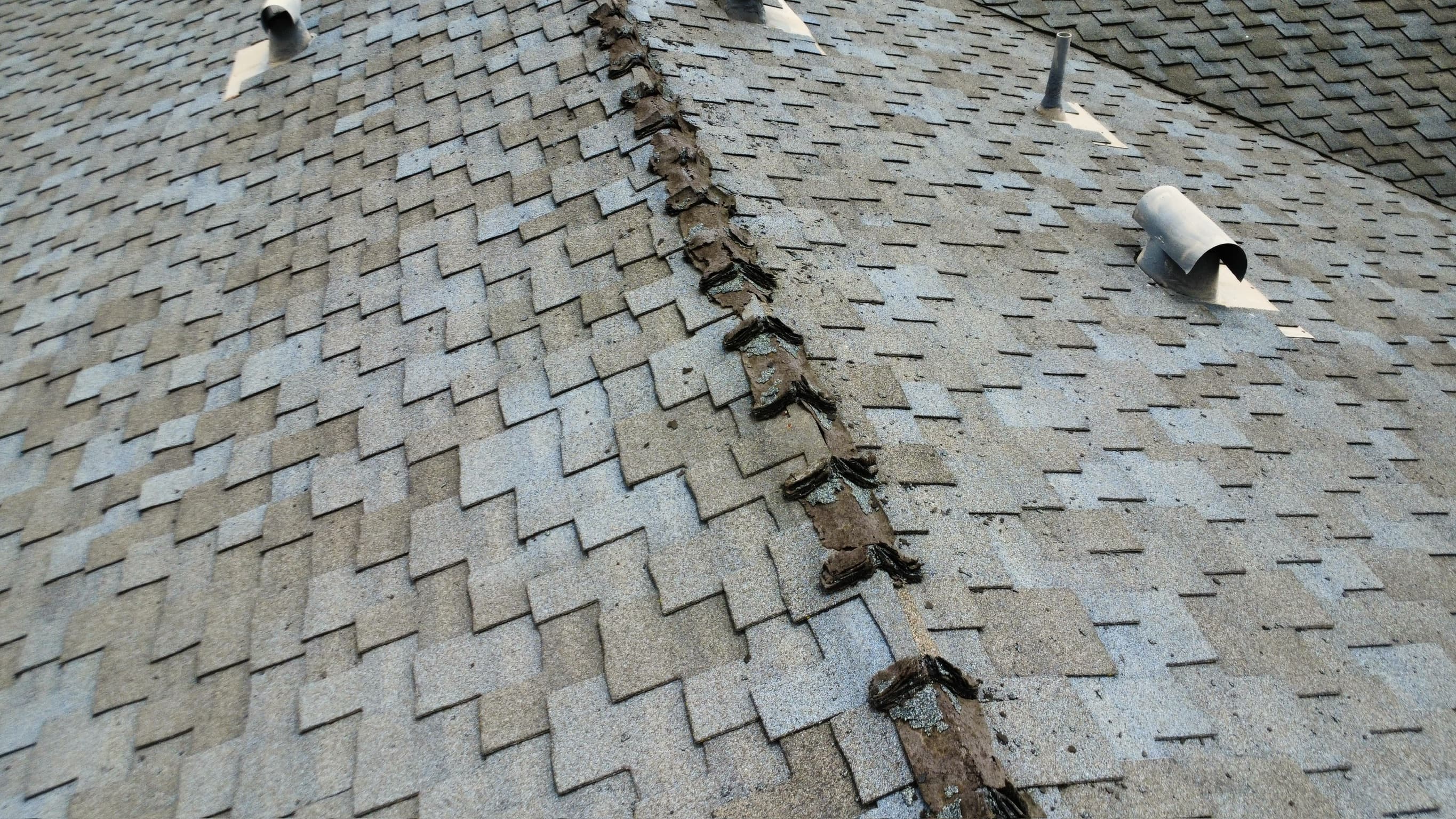 View down a roof ridge where every cap shingle has split into layers and lost its granules, leaving bare mat open to the sky, while the composition field on both slopes is still intact.