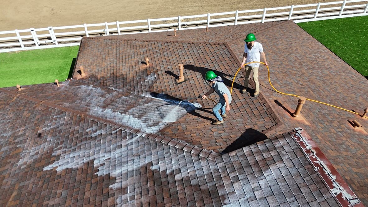 Aerial view of two crew members treating a hip roof, white spray visible off the wand and a milky arc of freshly sprayed Roof Maxx spreading across the brown shingle beside it