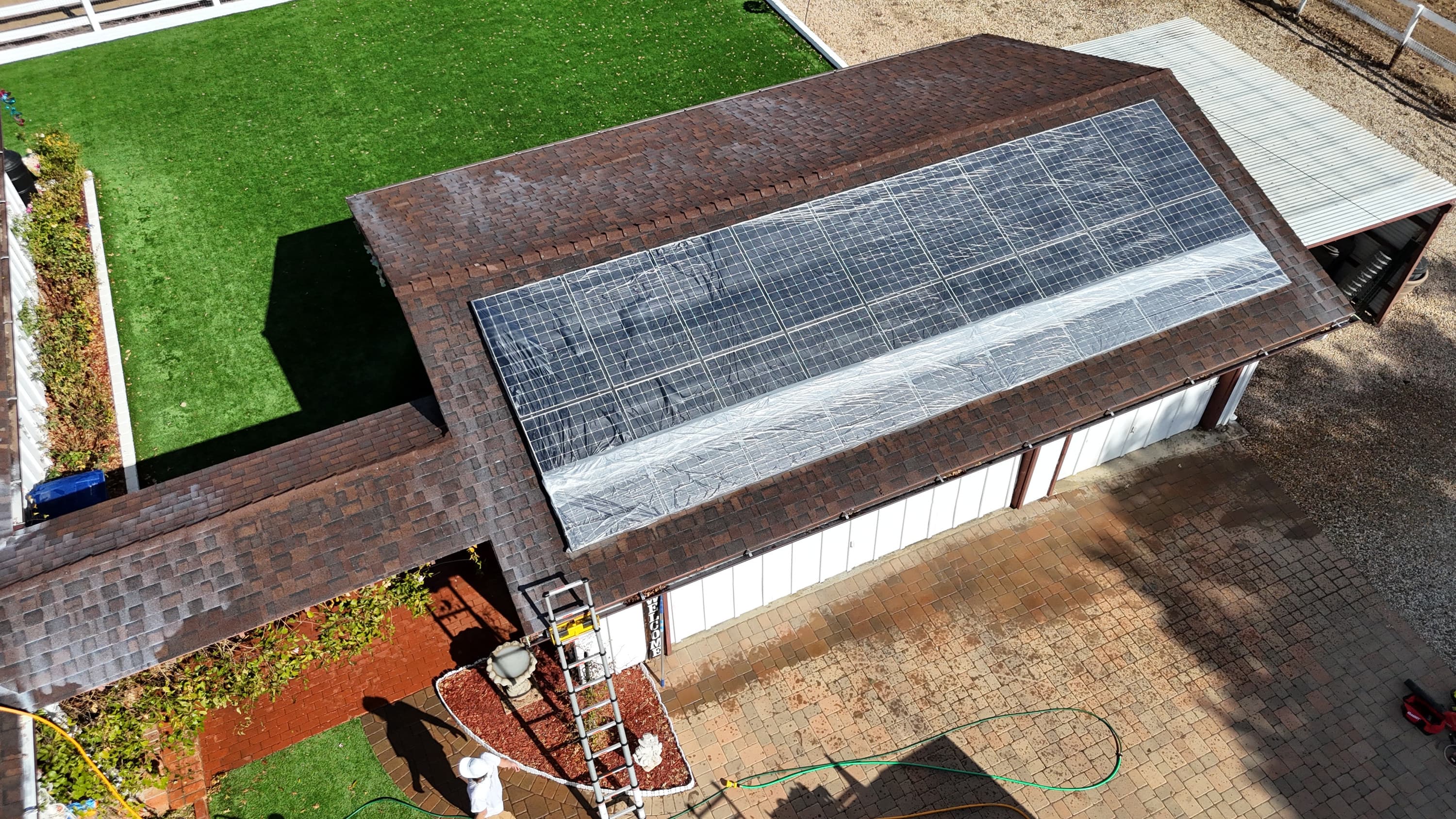A roof-mounted solar array covered edge to edge in protective sheeting, the shingle around it darkened where the treatment has soaked in, a ladder at the eave