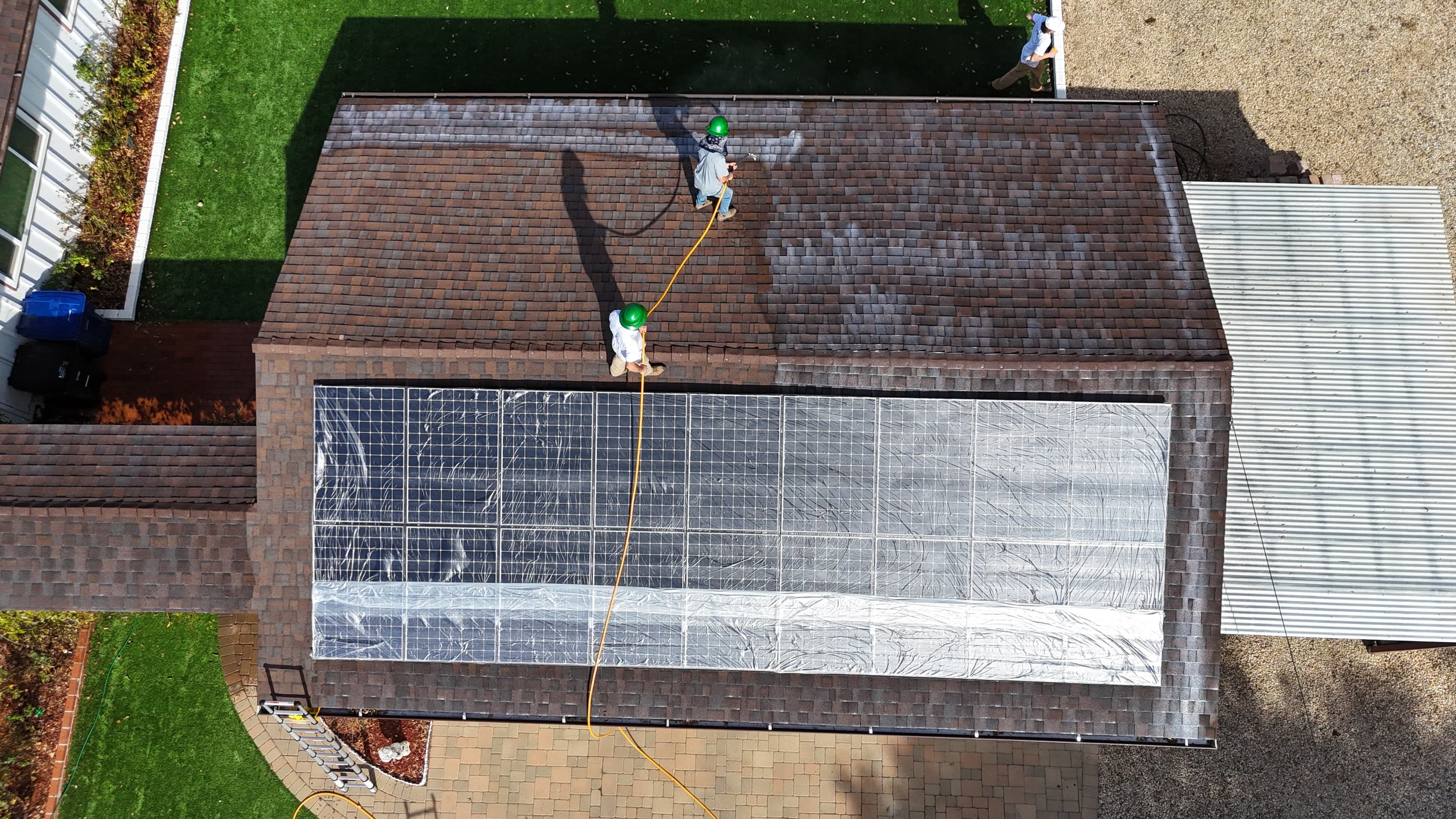 Overhead view of a solar array sheeted over in plastic while two crew members spray Roof Maxx across the shingle above it